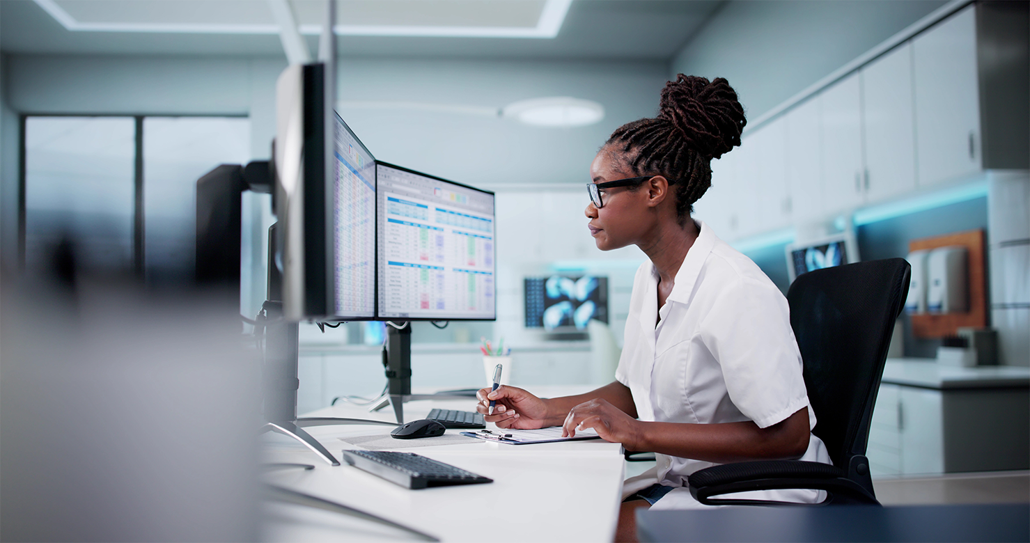 Young Black optometrist is facing her monitor that is displaying spreadsheets. She is focused and completing some calculations on a note pad.