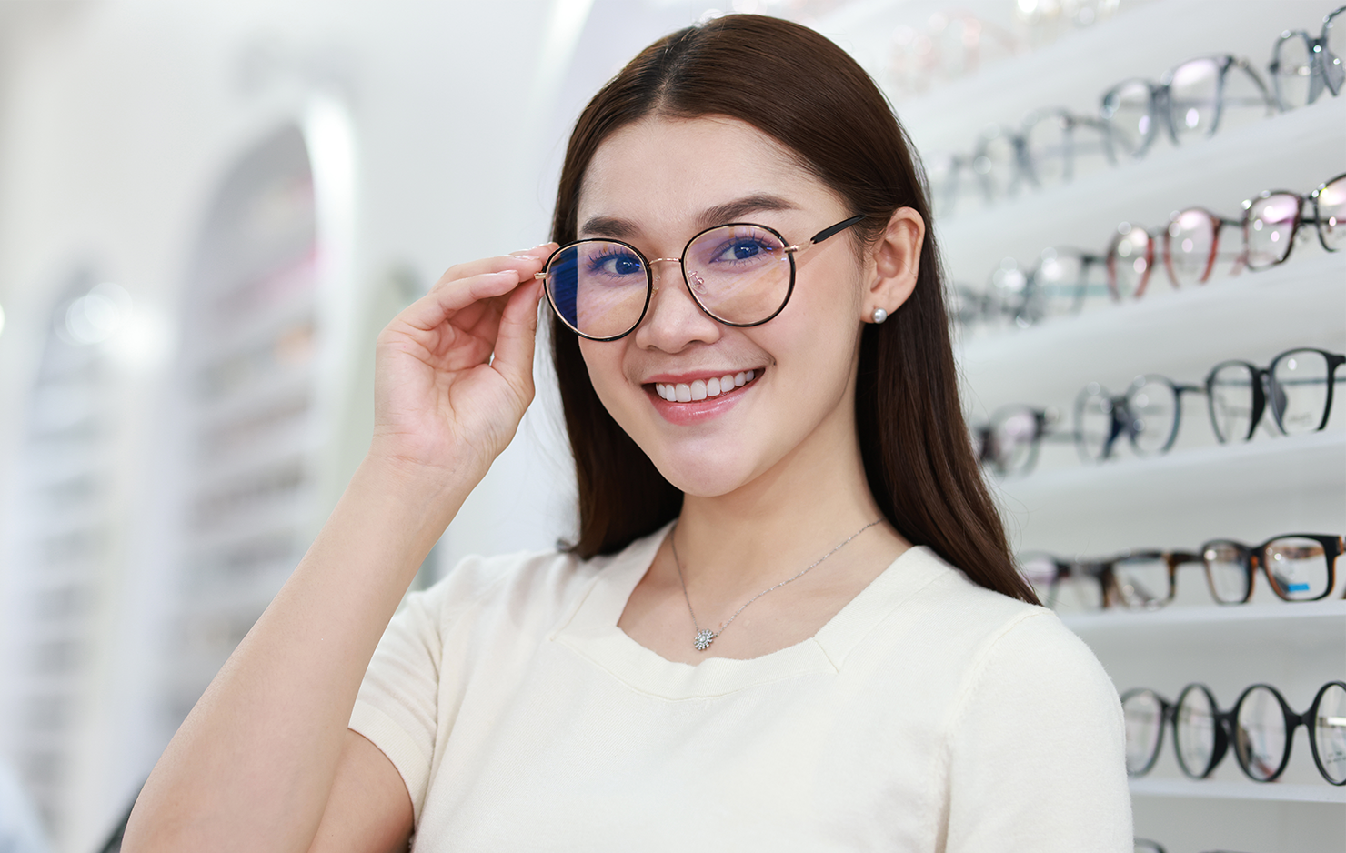 Young female optometrist is facing the camera, smiling. She is wearing round glasses and holding the right edge of the frame. She is standing in front of a wall of glasses.