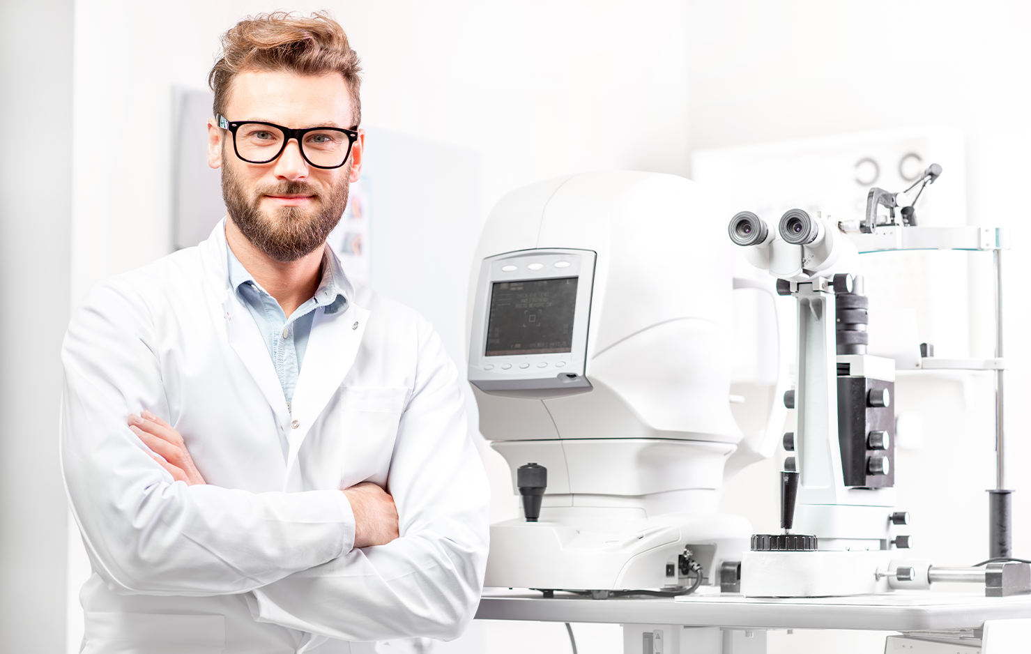 Male Optometrist facing the camera, wearing a white doctor's coat. Smiling with his arms folded. There is optometric equipment behind him.