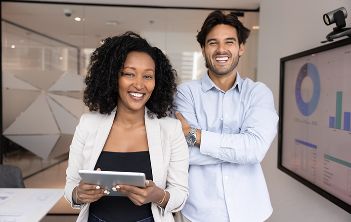 A female and male optometrists are facing the camera. The woman is Black and is holding a tablet, smiling. The man is brown with his arms folded, also smiling.