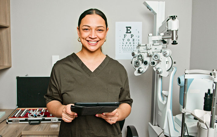 An optometrist wearing scrubs smiles, looking directly at the camera while holding a tablet used for advanced technology in eye exams. A phoropter, Snellen chart, and other examination equipment are in the background.